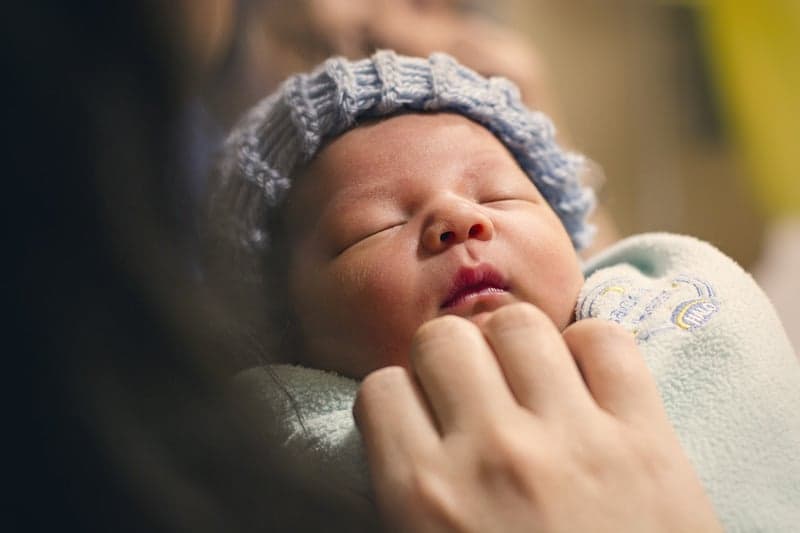 A sleeping newborn in a knit cap, held close in soft natural light